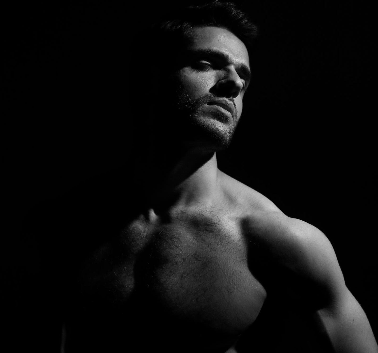 A black and white studio portrait of a muscular, shirtless man in dramatic side lighting against a solid black background.